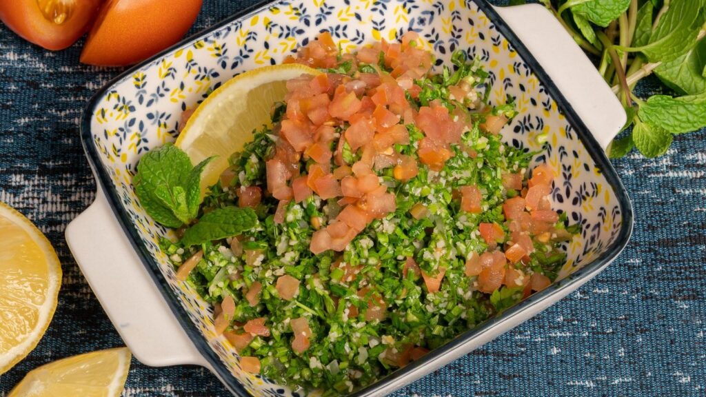 Taboulé vert servi dans un plat en céramique avec des tomates et un citron à côté